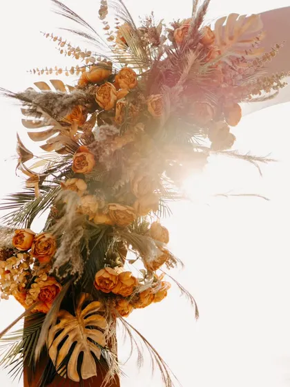 A close-up of a rustic floral arrangement featuring pampas grass and dried leaves. The sun flare adds a dreamy, golden-hour touch to this boho-chic decor detail.