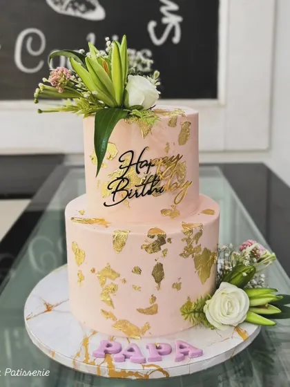 A beautiful two-tiered birthday cake for "Papa" in a soft pink hue. The cake is decorated with gold leaf, fresh white roses and lilies, and a "Happy Birthday" topper.