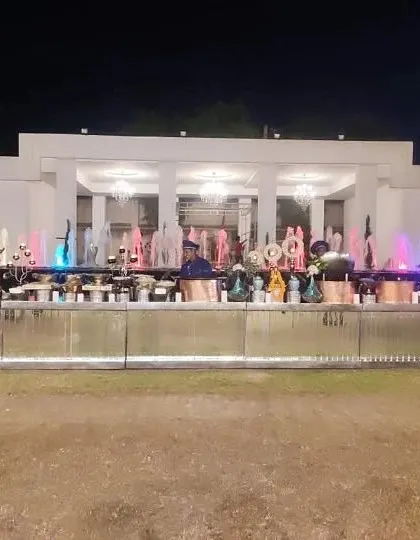 A spectacular night-time buffet setup in front of a fountain with colorful lights. The long, illuminated counter adds a modern and festive touch to this large-scale wedding reception.