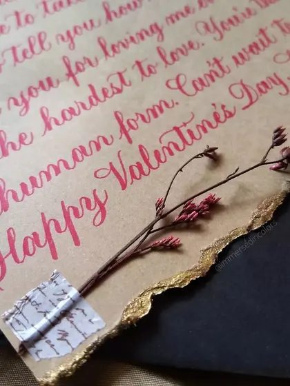 A close-up of a handwritten letter, showing the texture of the paper and the detail of the brush pen calligraphy. The little sprig of dried flower adds a delicate, natural touch.