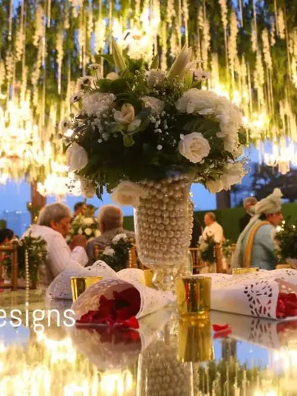 A close-up of a guest table under a canopy of hanging flowers and lights. The reflection on the mirrored tabletop creates a beautiful, layered effect, doubling the magic of the decor above.