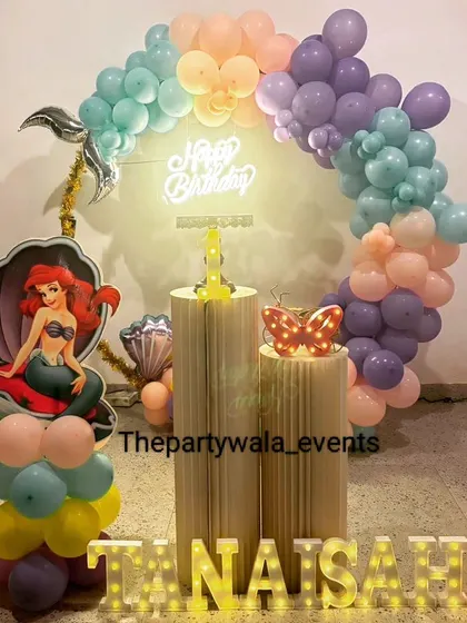 A mermaid theme with a pastel balloon arch and a 'Happy Birthday' neon sign. The Ariel cutout in a seashell is a great touch.