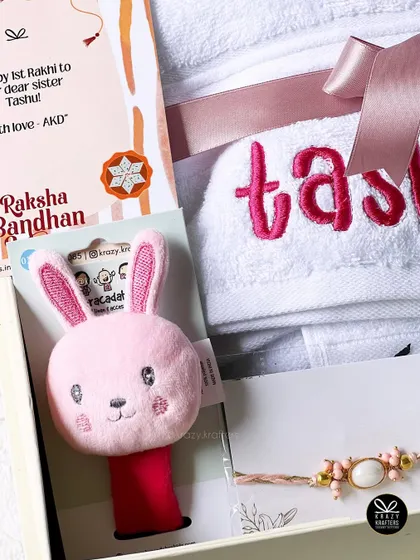 A sweet Rakhi hamper for a baby sister, featuring a personalized towel, a cute pink bunny wrist rattle, and a Raksha Bandhan card.