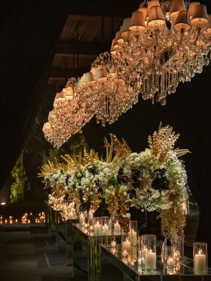 An awe-inspiring view of a long table at a black and gold cocktail event. Rows of crystal chandeliers hang above, their light reflecting off mirrored surfaces and illuminating the dramatic black, white, and gold floral arrangements.