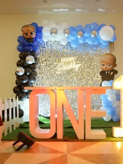 A Boss Baby first birthday setup with a silver shimmer wall and a "ONE" table. The decor includes a personalized milestone board.