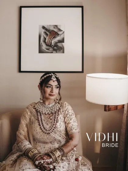 A sophisticated bridal portrait. The bride is seated indoors, with a framed photo and a modern lamp adding to the chic, editorial feel of the image.