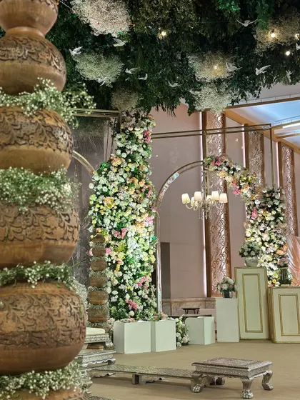 A detail shot of the vintage garden decor, featuring traditional carved pots and towering floral installations. The ceiling is a canopy of green leaves and hanging white birds.