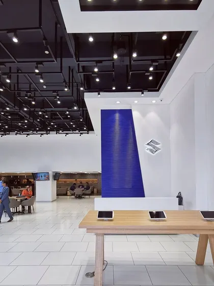 The digital interaction zone in a Maruti Suzuki Arena showroom, featuring tablets on a central wooden table. This area is defined by a bold blue accent wall and the iconic Suzuki logo, creating a focal point for brand engagement.