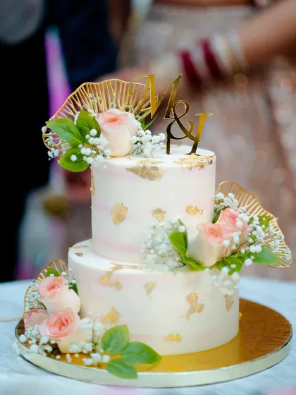 A romantic two-tiered cake featuring a soft pink and white watercolor effect, touches of gold leaf, and a custom "M&Y" monogram topper. The cake is beautifully finished with fresh pink roses and baby's breath.