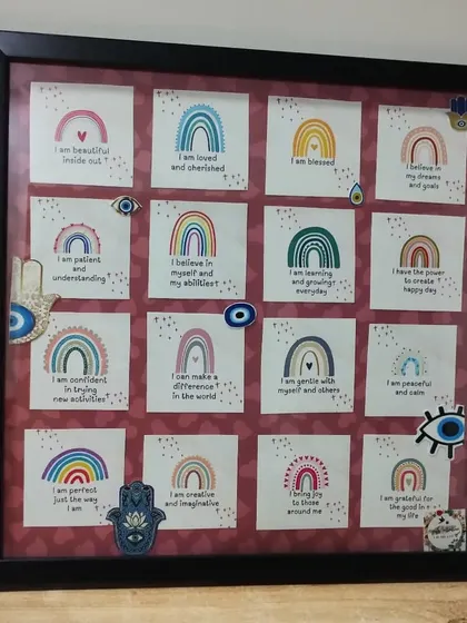 An affirmation board I created for actress Helly Shah, featuring positive "I am" statements on rainbow-themed cards. The frame is decorated with evil eye and hamsa hand charms for protection.
