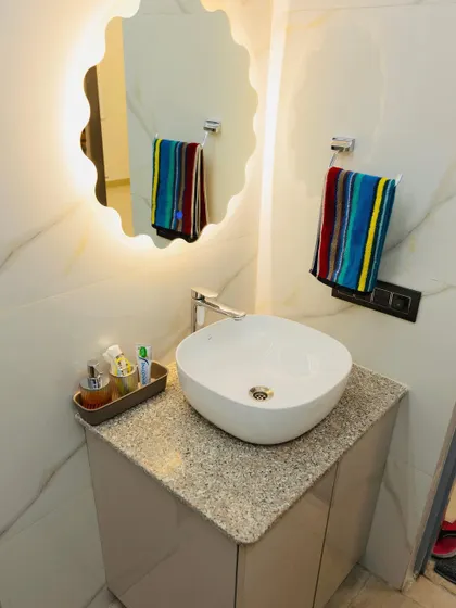 A close-up of a bathroom vanity area. The design features a stylish vessel sink, a granite countertop, and a unique flower-shaped backlit mirror that adds a touch of elegance.