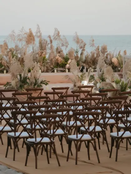 Rows of wooden cross-back chairs set up for a rustic and elegant beach wedding ceremony against a backdrop of pampas grass.