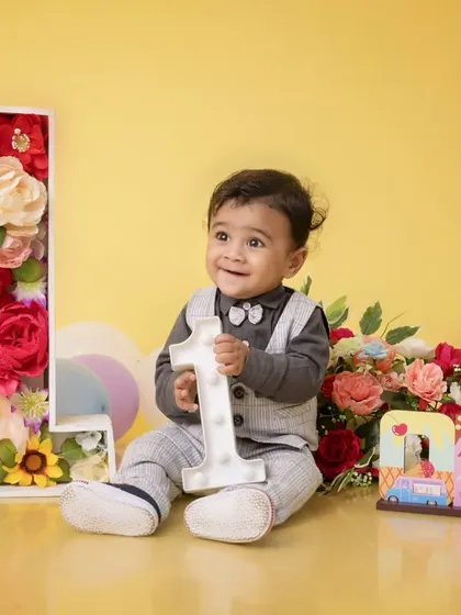 A beautiful first birthday setup with a floral number '1' and bouquets of flowers. The baby boy, dressed in a smart vest, holds a light-up number one, adding a touch of magic to the scene.