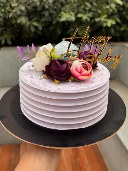 A lovely lavender cake with a ribbed buttercream finish. It's topped with a beautiful arrangement of artificial flowers in shades of purple and pink.