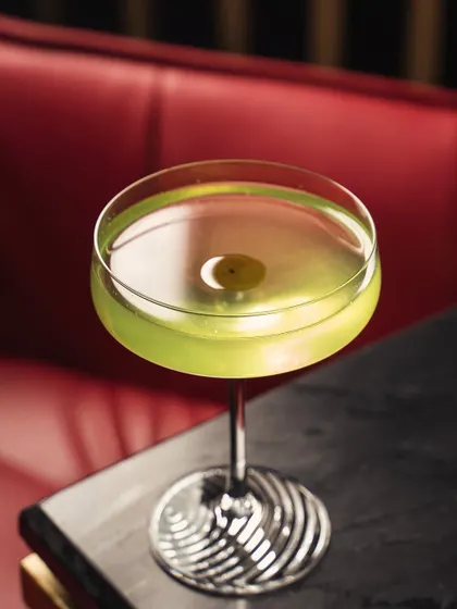 A sophisticated green cocktail in a coupe glass, shot against a rich, red leather booth. The composition and lighting create a classic, upscale speakeasy mood.