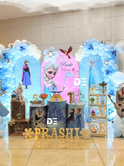 An elegant 'Frozen' theme for Prashi's 7th birthday. The setup features two blue balloon arches, cutouts of Elsa and Olaf, and a winter castle theme.