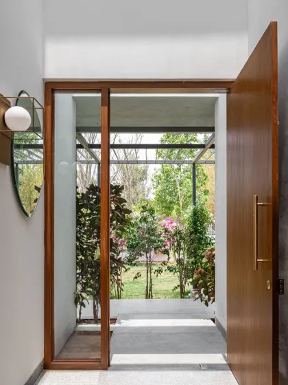 The main entrance opens to a shaded walkway and patio, immediately establishing a connection with the outdoors. The clear glass and wood framing provide a transparent threshold between the garden and the interior foyer.