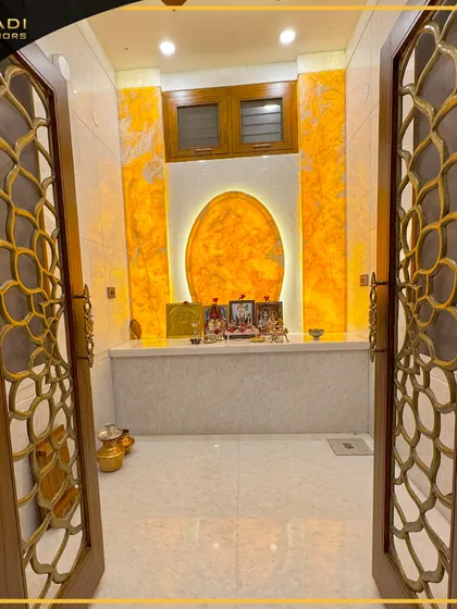 A view into a stunning puja room through intricately designed doors with a floral metal jali pattern. The back wall glows with a backlit onyx panel shaped like the sun, creating a divine ambiance.