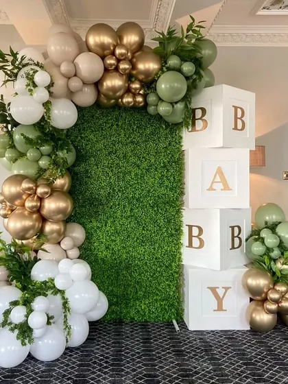 A baby shower setup featuring a green hedge wall with a gold and green balloon arch. The "BABY" blocks add a classic touch to this garden-inspired theme.