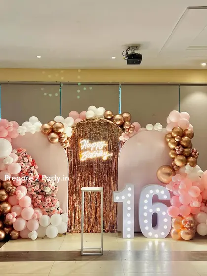 A pretty in pink sweet sixteen birthday decoration. This large-scale setup features double-arched backdrops, a rose gold shimmer panel, a neon sign, and an extensive balloon garland in shades of pink, white, and rose gold.