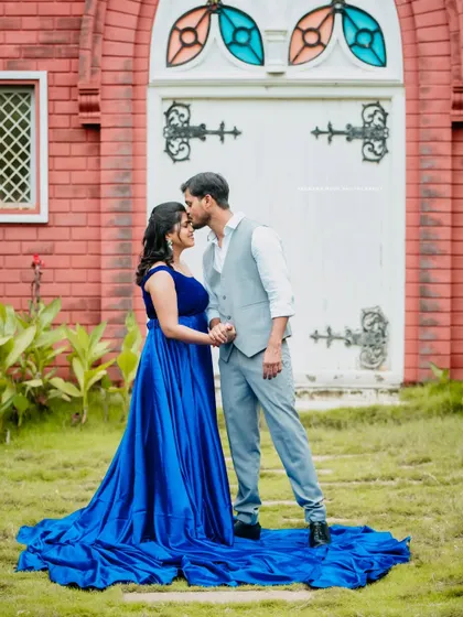A tender moment as the partner kisses the mother-to-be's forehead. They stand in a beautiful blue gown before a charming building, creating a fairytale-like scene.
