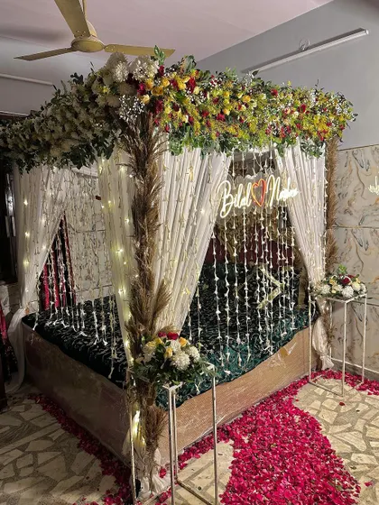 A rustic and elegant wedding night setup. The bed is decorated with a floral canopy featuring pampas grass and a custom neon sign.