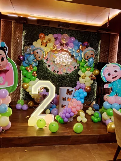 A Cocomelon-themed second birthday with a circular balloon arch, character cutouts, and a light-up number '2' set against a leafy green wall.