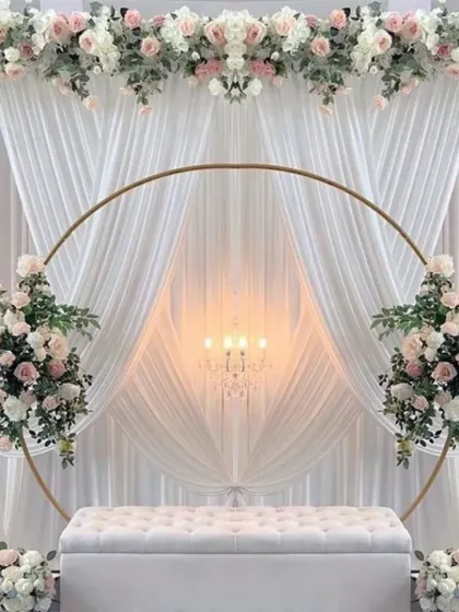 An absolutely breathtaking wedding or engagement stage. The setup features a large golden ring, elegant white draping, and beautiful arrangements of white and pink artificial flowers.