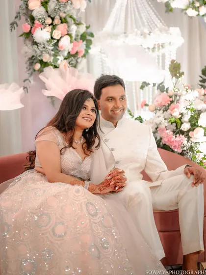 The couple seated together against a beautiful backdrop of white and pink florals. A lovely portrait from their fun filled ring ceremony.