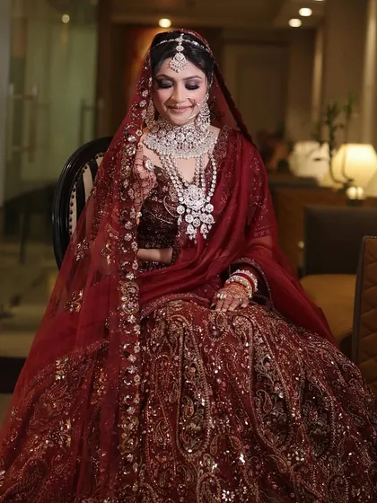 A classic bridal portrait, seated and smiling. The bride's magnificent maroon velvet lehenga and diamond jewelry are on full display, capturing a moment of quiet joy.