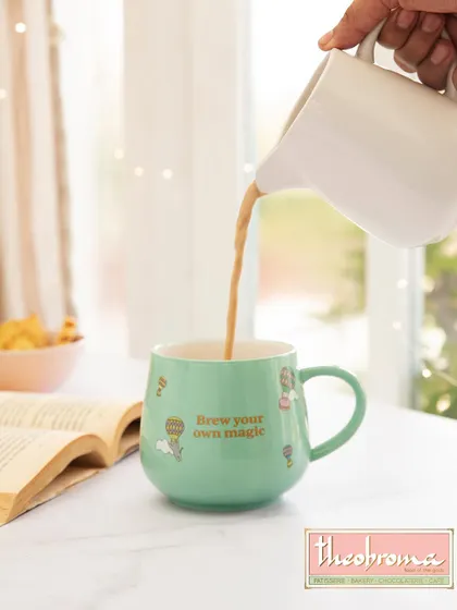 A lifestyle product shot for Theobroma, capturing a quiet moment of indulgence. The action of pouring tea into the branded mug, set against a soft, book-filled background, creates a cozy and aspirational scene.