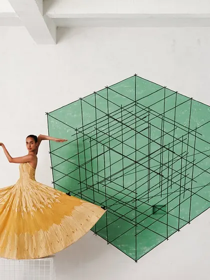 A model in a yellow Banarasi drape appears to float in front of a green geometric installation, creating a surreal and colorful composition.