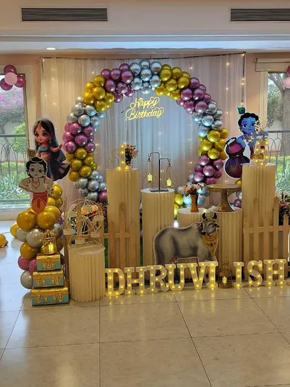 A Krishna-themed party in a well-lit room with large windows. The setup includes a balloon arch, character cutouts, and marquee letters.