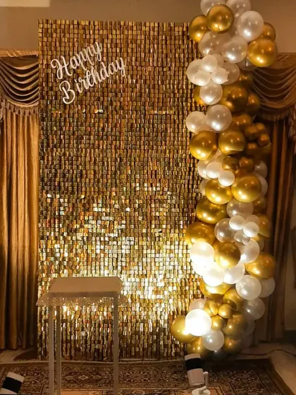 An elegant birthday photo corner at home. A gold sequin wall provides the shimmer, complemented by a cascade of white and gold balloons and a simple 'Happy Birthday' sign.