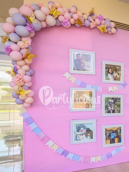 A lovely memory wall with framed photos, a pastel banner, and a delicate balloon garland with butterfly accents.