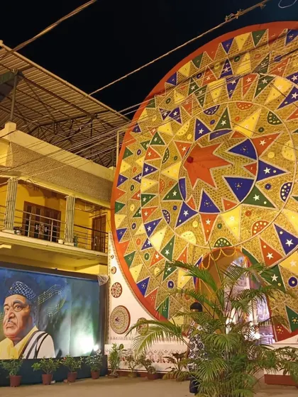 An exterior night view of the Durga Puja pandal, featuring a massive, colorful 'Japi' (traditional Assamese hat) and a mural of Bhupen Hazarika.