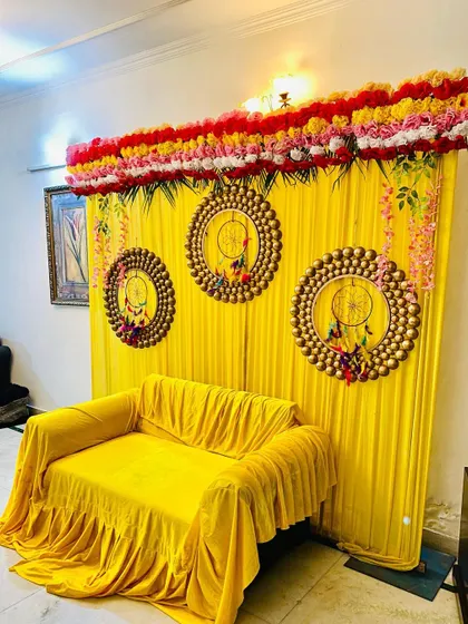 For a home mehndi or haldi function, I can create a vibrant photo corner like this one. This setup includes a bright yellow backdrop with matching sofa covers, decorative dreamcatcher-style hangings, and a floral border on top.