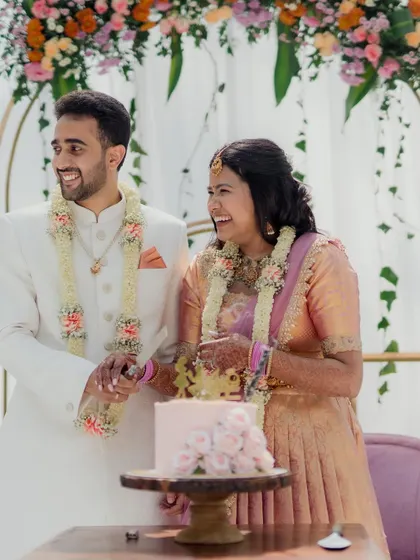 A joyous cake-cutting moment at an engagement ceremony. The couple is all smiles, seated on a modern pink sofa against a backdrop of golden arches and pastel floral decor.