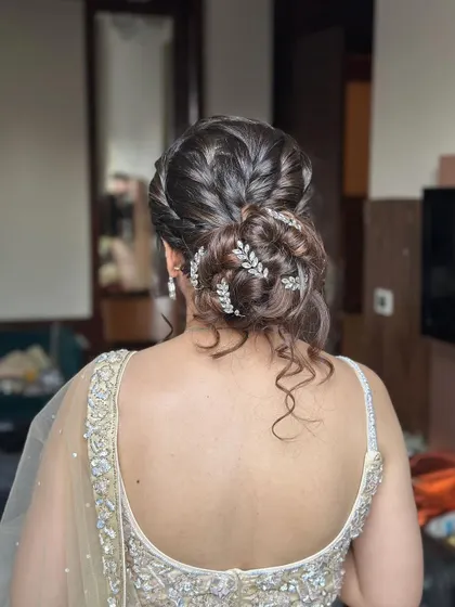 This messy bun features interwoven braids and is decorated with delicate silver leaf accessories. The loose curls around the face add a touch of softness, making it ideal for an engagement or reception.