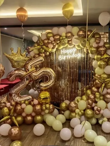 A lavish 25th birthday setup with a gold fringe backdrop, a massive arrangement of gold and white chrome balloons, and a large crown prop.