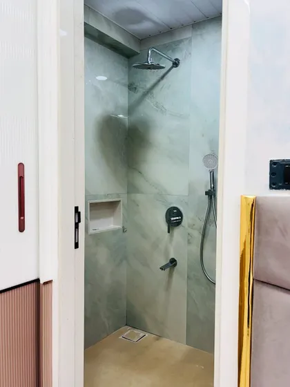 A view into the shower area, which is separated by a frameless glass door. The walls are clad in light green marble-finish tiles, creating a serene and calming atmosphere.