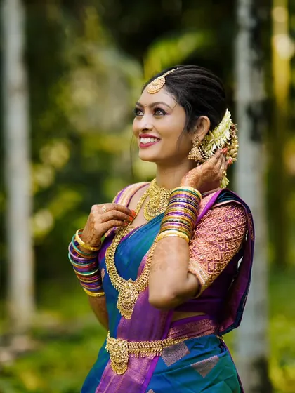 A candid bridal portrait taken outdoors, with the bride smiling as she adjusts her earring, looking radiant in her blue and purple saree.