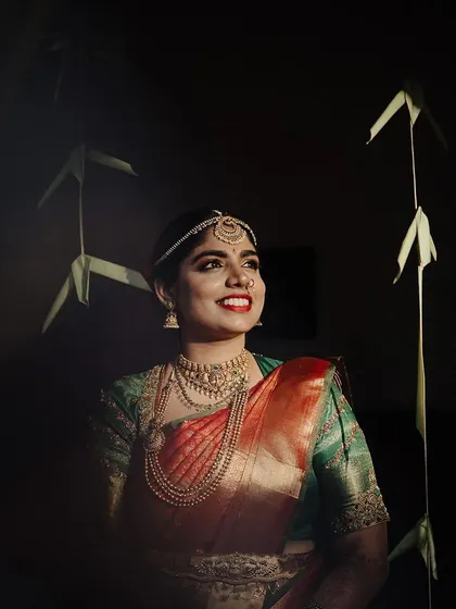An artistic bridal portrait using dramatic lighting. The bride is illuminated against a dark background, creating a spotlight effect that highlights her smile and traditional jewelry.