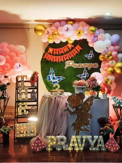 A whimsical fairy and butterfly theme for Pravya's birthday. The decor features a grass backdrop, a glittery fairy cutout, and a beautiful pink and gold balloon arrangement.