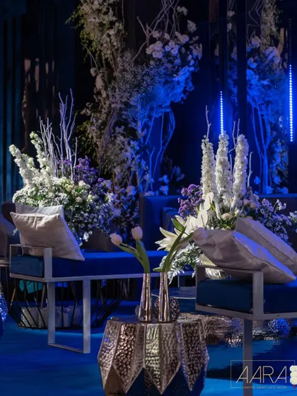 A seating arrangement from the 'Gala Galaxy Night', featuring modern blue velvet benches and silver accent tables. The space is filled with white and purple floral arrangements, contrasting beautifully with the deep blue lighting.