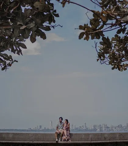 A wide, cinematic shot of Ashleen and Ranjodh sitting together on the Marine Drive promenade, with the famous Queen's Necklace skyline of Mumbai stretching out behind them.
