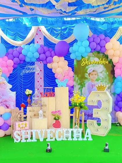 A vibrant outdoor birthday celebration under a tent. The colorful balloon arch and custom backdrop create a festive atmosphere for a third birthday party.