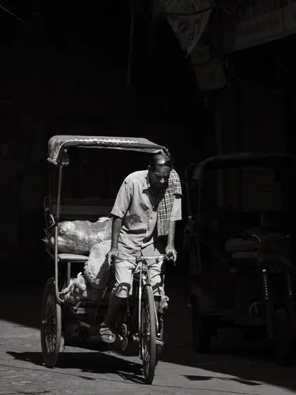 A cycle-rickshaw driver pedals through a dark lane, his form highlighted by a sliver of light. This black and white image captures the daily toil and quiet dignity of workers in Old Delhi.
