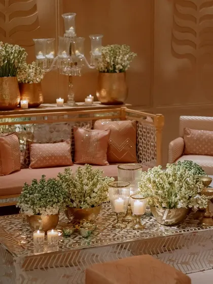 A luxurious lounge area with plush sofas and a mirrored coffee table. The space is filled with large brass pots of white flowers and elegant glass candelabras.