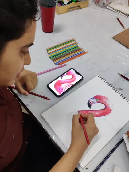 A student uses a reference on her phone to carefully draw a flamingo, focusing on capturing the bird's elegant curves with colored pencils.